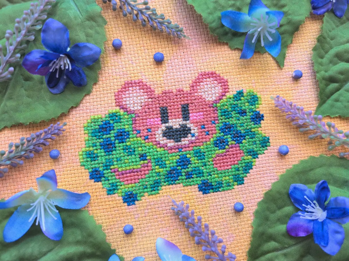 A cross stitch on mottled orange aida of a pastel coloured bear sticking out of a blueberry bush, his mouth covered in blueberry stains. The cross stitch is framed by green plastic leaves and blue plastic flowers.