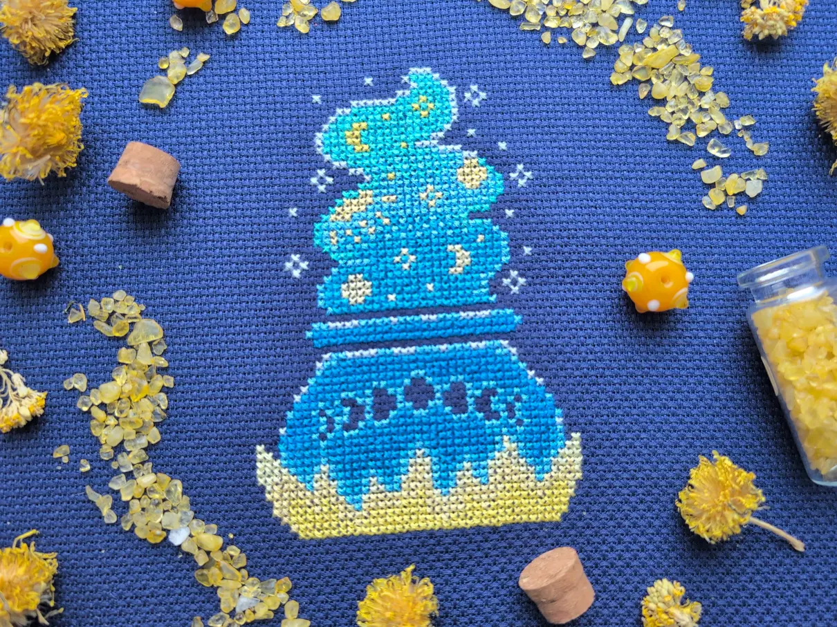 A cross stitch on navy aida of a blue ombre cauldron. Bubbling out of the cauldron is a blue ombre gradient stream plume with yellow suns, moons, and stars scattered throughout it.