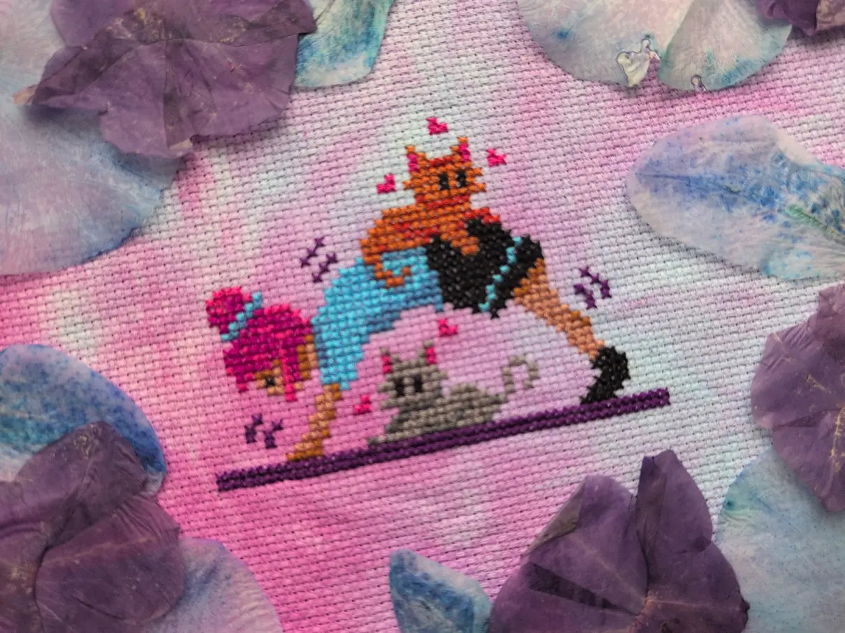 A cross stitch on mottled blue and purple aida of a person doing the downward dog yoga pose with one cat sitting under them and another on their back. The cross stitch is framed with dried blue orchid petals and pressed purple petunia blooms.