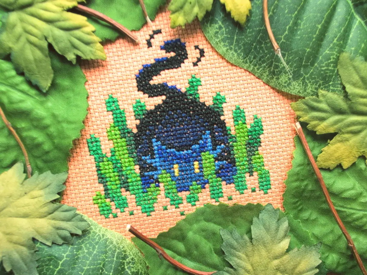 A cross stitch on orange aida of a cat crouched down in a patch of grass thinking he is well hidden though he is not. The cross stitch is framed by various plastic leaves and twigs.