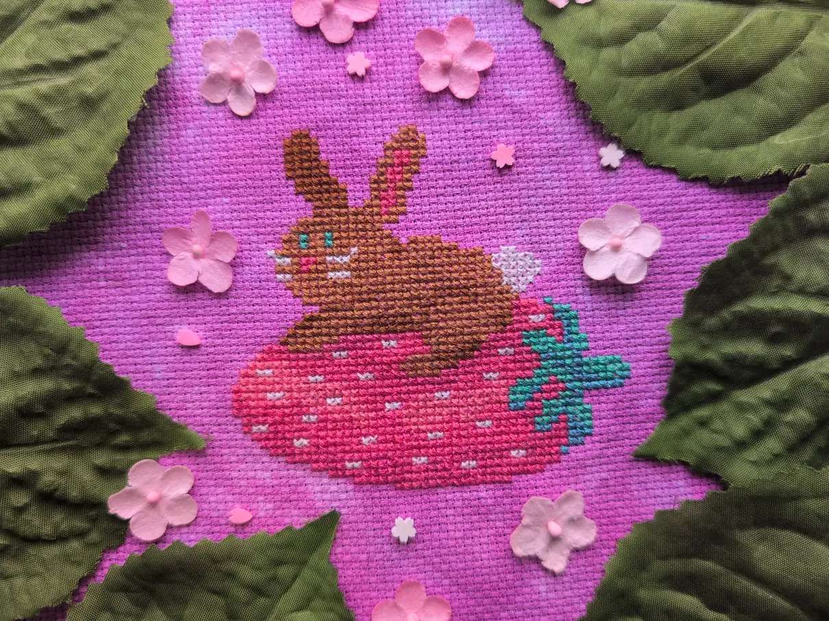 A cross stitch of a brown bunny with a white tail and whiskers perched atop a strawberry. The cross stitch is framed by fake green leaves and pink and white paper flowers.