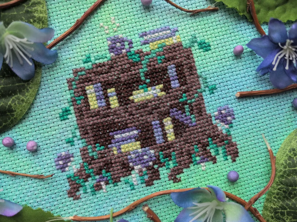 A cross stitch of a bookshelf carved directly out of a large tree stump on green and blue aida. Several books are on the shelves, mushrooms and leafy vines surround the bookshelf, and a steaming purple mug sits on top. The cross stitch is framed by plastic twigs, leaves, and blue flowers.