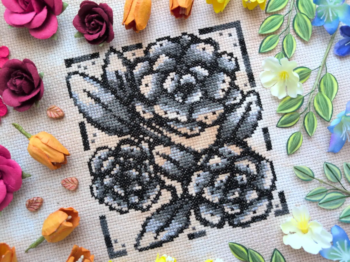 A greyscale cross stitch on tea-dyed aida of three sketched flower blooms. It's framed with many different flowers and leaves arranged in the colours of the rainbows.