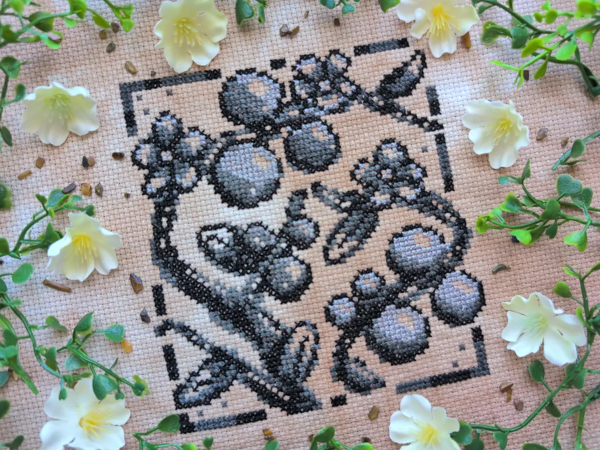 A greyscale cross stitch on tea-dyed aida of sketched tomatoes and flowers on a vine climbing upwards. It's framed with white flowers and green vines.
