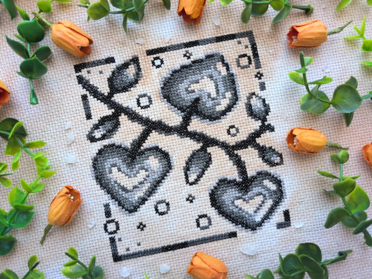A greyscale cross stitch on tea-dyed aida of a climbing vine with heart shaped fruits. It's framed with orange flowers and green vines.