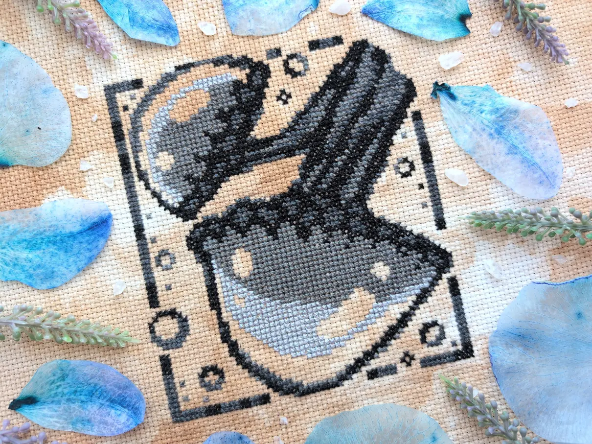 A greyscale cross stitch on tea-dyed aida of sketched glowing mushrooms. It's framed with translucent blue orchid petals and white pebbles.