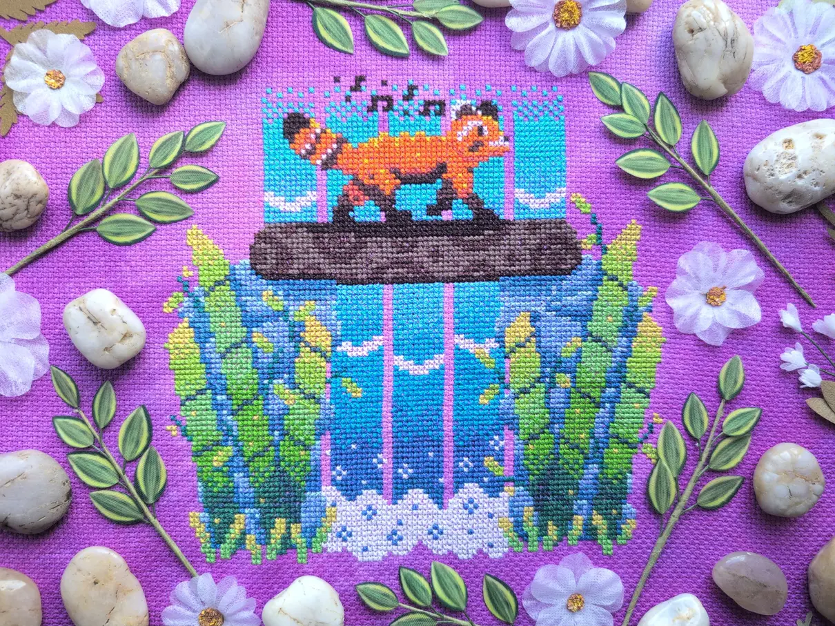 A cross stitch on purple and pink mottled aida of a red panda crossing a log over a waterfall. Bamboo plants sprout from the bottom of the waterfall. The cross stitch is framed with smooth beige rocks, fabric leaves and branches, and white fabric flowers.