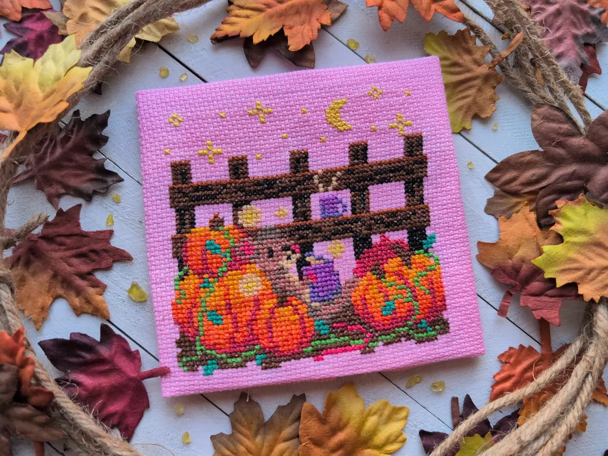 A cross stitch on pink aida of a mouse reading a book illuminated by fireflies nestled in a pumpkin patch. The cross stitch is framed by rough, fibrous rope and fall-coloured leaves.