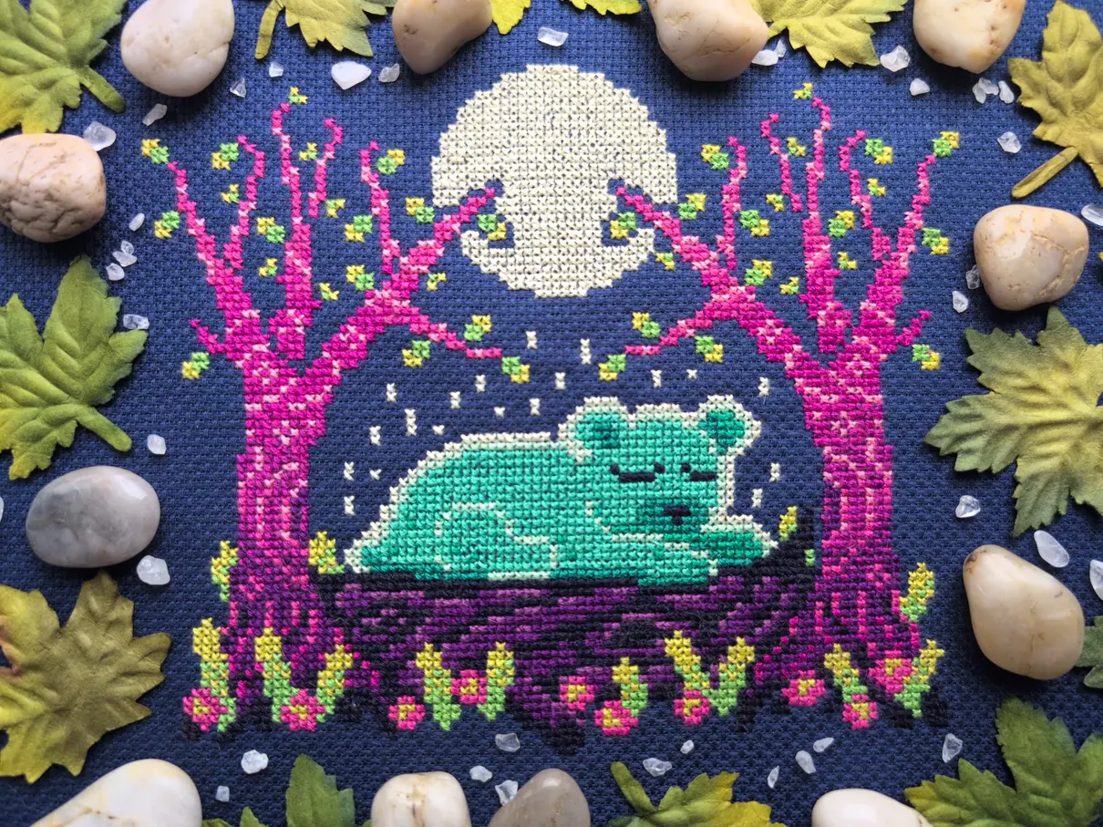 A cross stitch on navy blue aida of a ghostly bear sleeping on a log between two purple ombre trees. The cross stitch is framed with green maple leaves and white rocks.