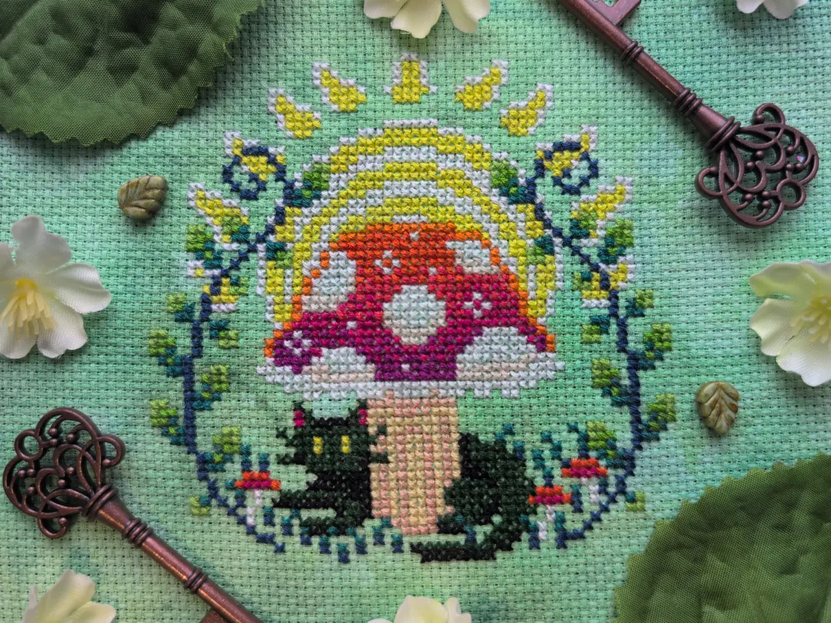 A cross stitch of a black cat sitting under a mushroom. Leafy plants surround the mushroom, and a sun is rising up in the background. The mushroom cap resembles the colours of a sunrise. 