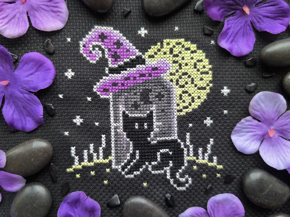 A cross stitch on black aida of a cat sitting in front of a grave stone. Atop the gravestone is a purple witch's hat and a large full moon shines behind them. The cross stitch is surrounded by fake purple flowers and black stones.