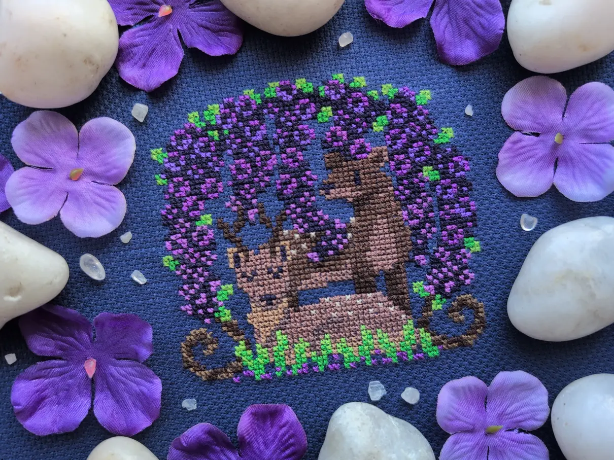 A cross stitch on navy blue aida of two deer resting under and arch of wisteria flowers. The cross stitch is framed by decorative white stones, translucent white pebbles, and purple flowers.